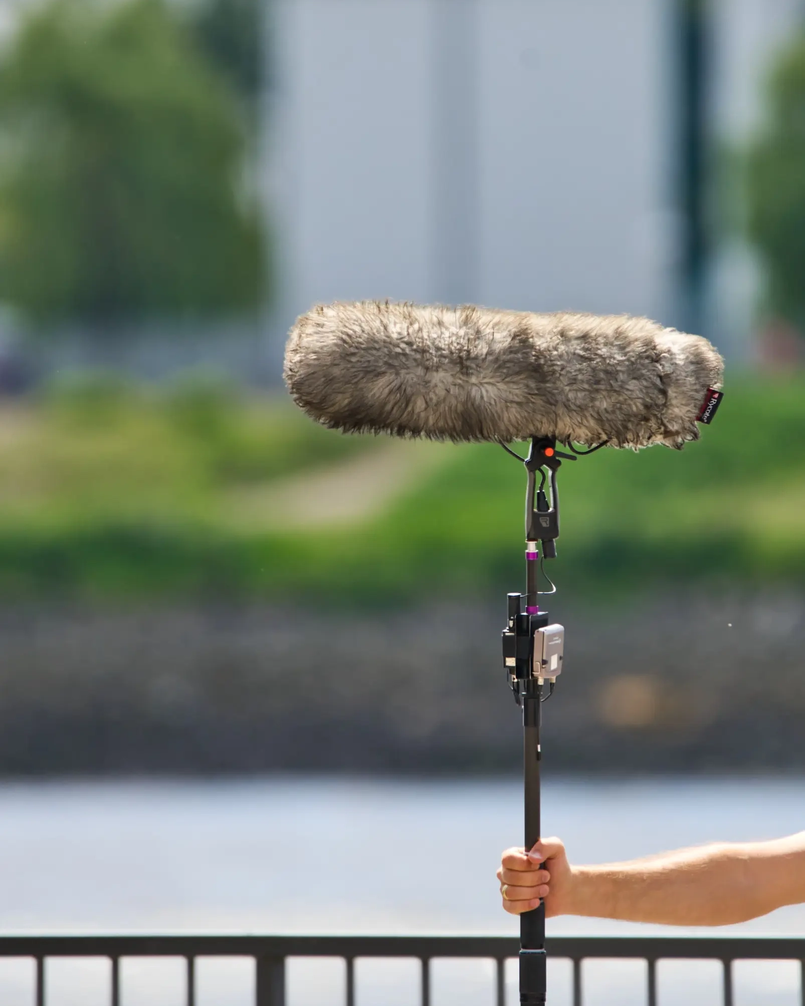 Una mano tiene un microfono a boom con un paravento peloso, sullo sfondo sfocato di un corso d'acqua.